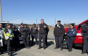 A group of emergency responders in uniform listen to a man speaking outdoors next to emergency vehicles on a clear, sunny day. Most are standing with arms crossed or hands in pockets.