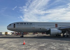 A large jet airplane with "WORLD" written on the fuselage is parked on a gravel area, surrounded by emergency vehicles and personnel under a partly cloudy sky. Orange cones are placed nearby.