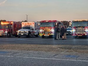 Several fire trucks and rescue vehicles are parked in a lot at dusk. Four people stand talking in front of the vehicles. The sky is clear and there are trees in the background.